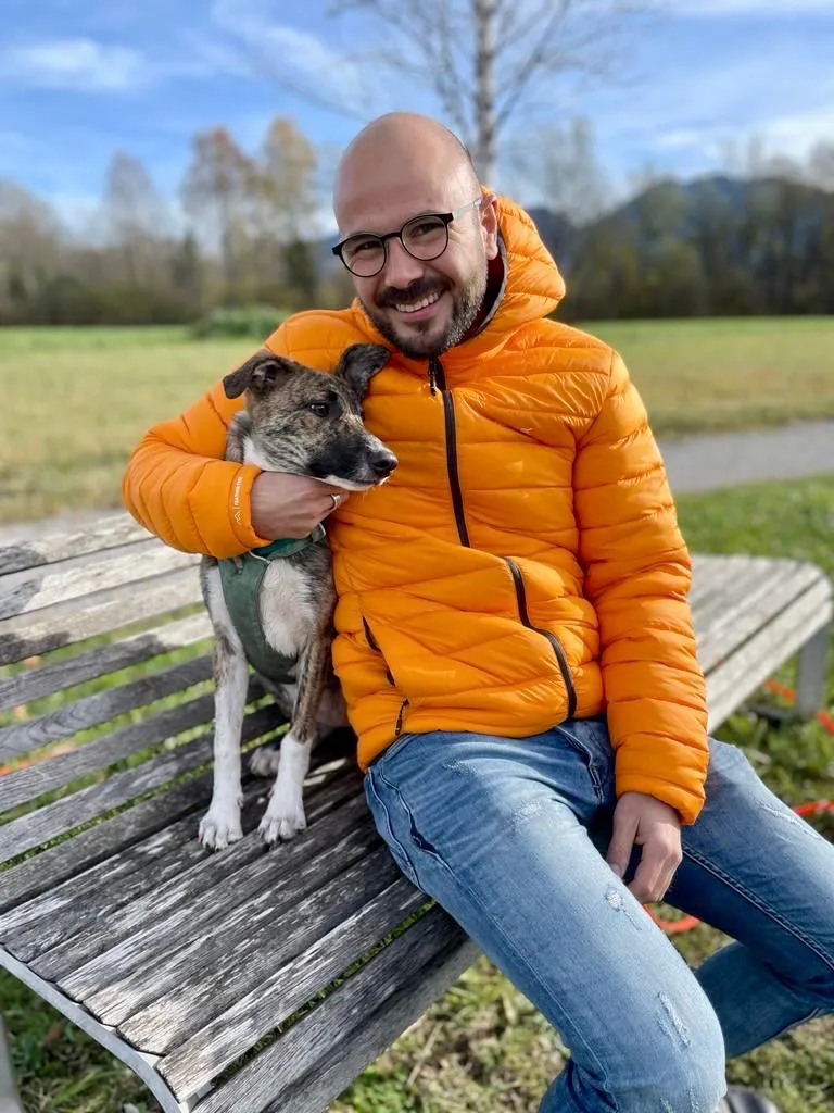 Simon, der mit seinem Hund auf einer Parkbank sitzt. Er lächelt in die Kamera und trägt eine grell orangene Jacke und eine Jeans.